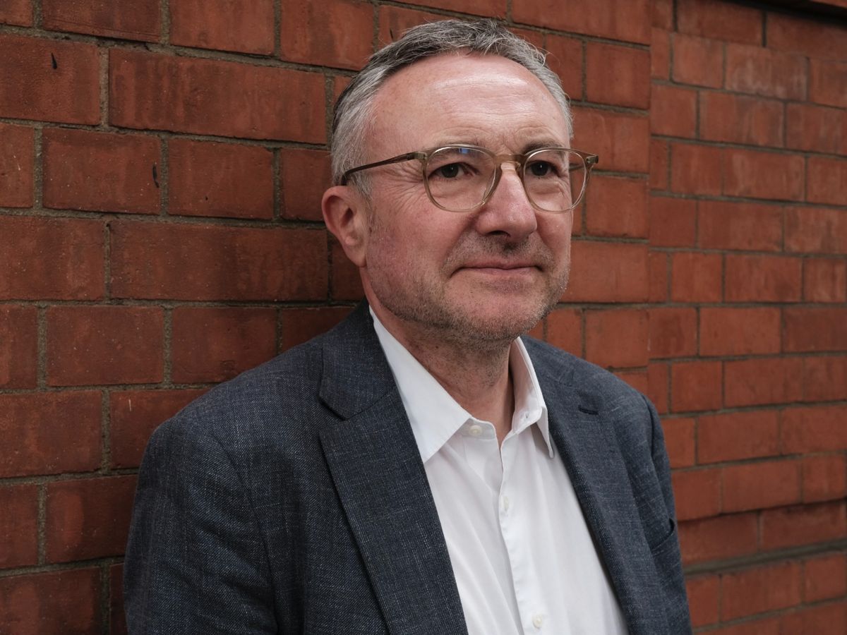 Professor David Owens, wearing a grey suit jacket and white shirt, stands in front of a brick wall. 